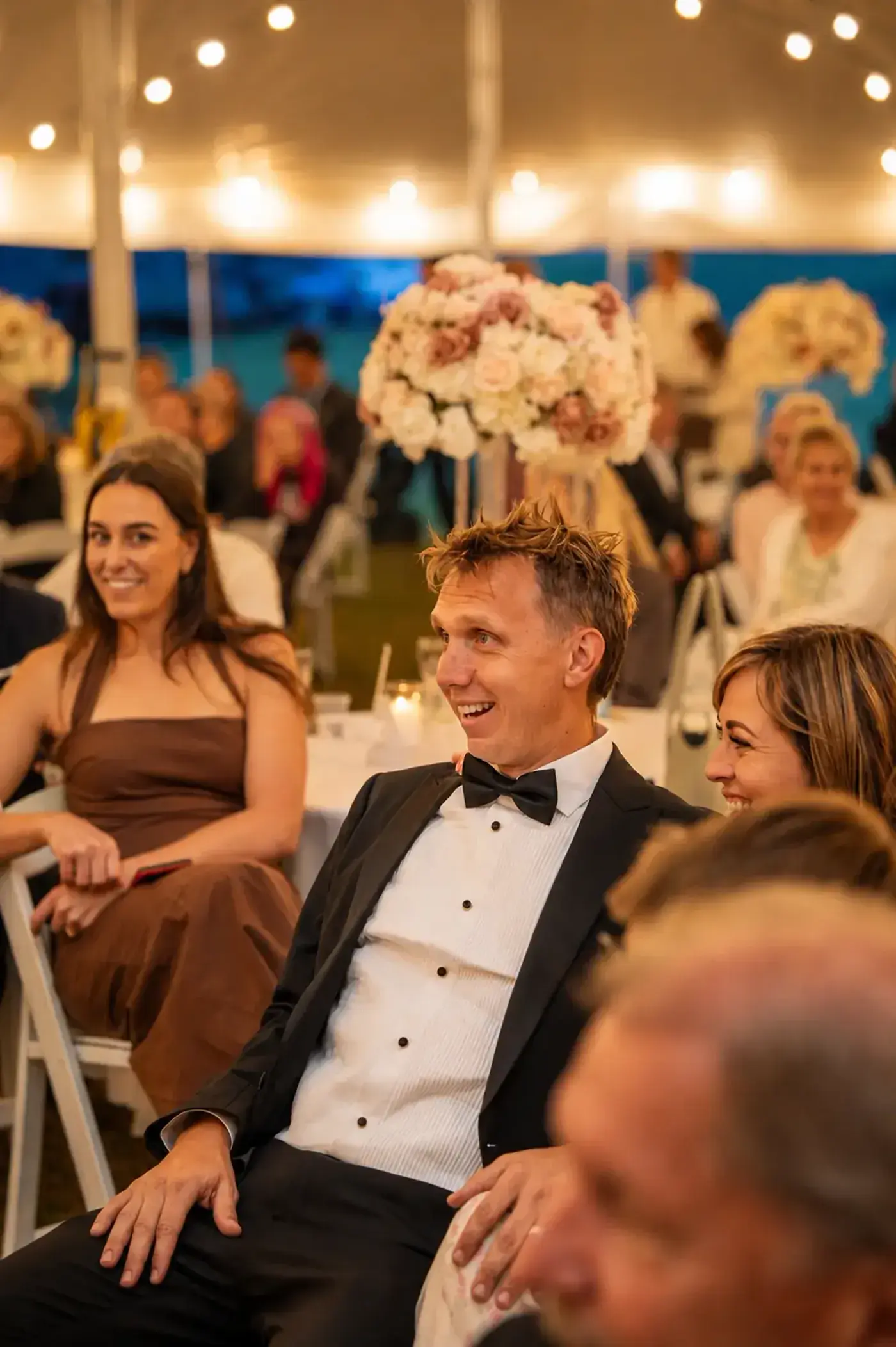 Wedding guests seated under festoon lights and tall floral centrepieces during a marquee reception at Adeline Estate